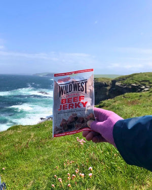 Hånd holder Wild West Beef Jerky Original ved kysten med havet i bakgrunnen