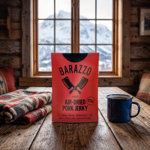 Barazzo Tørket Svinefilet Klassisk på et trebord i en hytte, med ullpledd, emaljekopp og snødekte fjell utenfor vinduet, AI-generert bilde