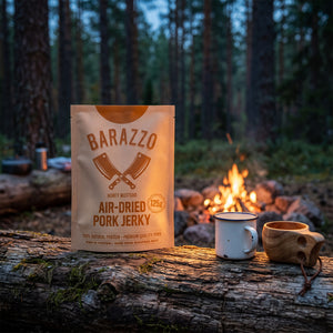 Barazzo Tørket Svinefilet Honning & Sennep på en stokk ved bålet i furuskogen i skumringen, med emaljekopp og kuksa, AI-generert bilde