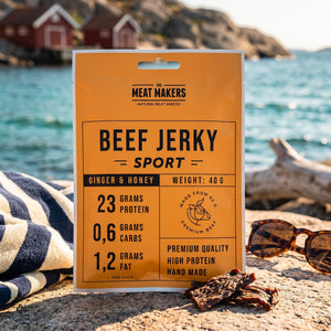 Meat Makers Sports Okse med Ingefær & Honning på et svaberg ved sjøen, med håndkle og solbriller, AI-generert bilde