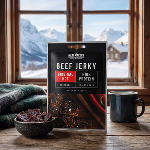 Meat Makers Beef Jerky Hot på et trebord i en hytte, med ullpledd, emaljekopp og snødekte fjell utenfor vinduet, AI-generert bilde