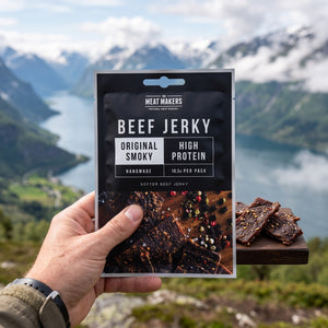Hånd som holder Meat Makers Beef Jerky Smoky foran et fjordlandskap, AI-generert bilde