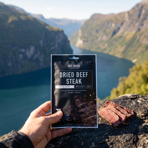 Hånd som holder Meat Makers Tørket Oksefilet foran et fjordlandskap, AI-generert bilde