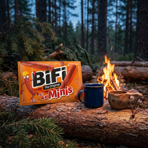 BiFi Minis 4-pack på en stokk ved bålet i furuskogen i skumringen, med emaljekopp og kuksa, AI-generert bilde