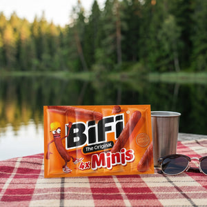 BiFi Minis 4-pack på et rutete piknikteppe ved et skogsvann en sommerdag, med stålkopp og solbriller, AI-generert bilde