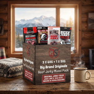 Big Brand Originals Beef Jerky Mixed Pack på et trebord i en hytte, med ullpledd, emaljekopp og snødekte fjell utenfor vinduet, AI-generert bilde