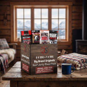 Big Brand Originals Beef Jerky Mixed Pack på et trebord i en hytte, med ullpledd, emaljekopp og snødekte fjell utenfor vinduet, AI-generert bilde