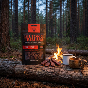 Biltong Premium Piri Piri på en stokk ved bålet i furuskogen i skumringen, med emaljekopp og kuksa, AI-generert bilde