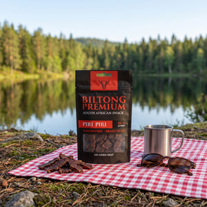 Biltong Premium Piri Piri på et rutete piknikteppe ved et skogsvann en sommerdag, med stålkopp og solbriller, AI-generert bilde
