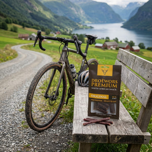 Biltong Premium Original Ølpølse (Droëwors) på en benk ved grusveien, med sykkel og sykkelhjelm, AI-generert bilde