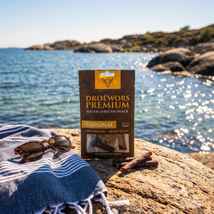 Biltong Premium Original Ølpølse (Droëwors) på et svaberg ved sjøen, med håndkle og solbriller, AI-generert bilde