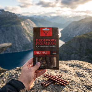Hånd som holder Biltong Premium Piri Piri Ølpølse (Droëwors) foran et fjordlandskap, AI-generert bilde