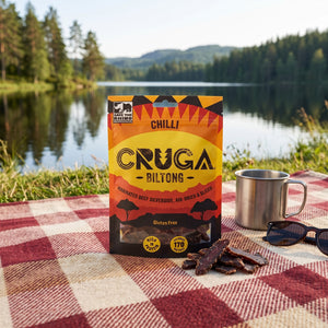 Cruga Biltong Chili på et rutete piknikteppe ved et skogsvann en sommerdag, med stålkopp og solbriller, AI-generert bilde