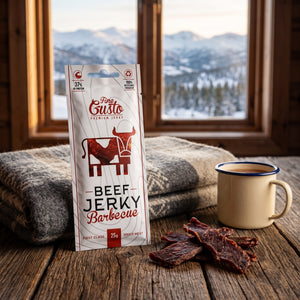 Fine Gusto Beef Jerky BBQ på et trebord i en hytte, med ullpledd, emaljekopp og snødekte fjell utenfor vinduet, AI-generert bilde
