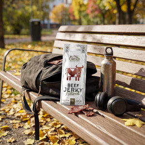 Fine Gusto Beef Jerky Naturell stikker opp av en ryggsekk på en parkbenk om høsten, med drikkeflaske og hodetelefoner, AI-generert bilde