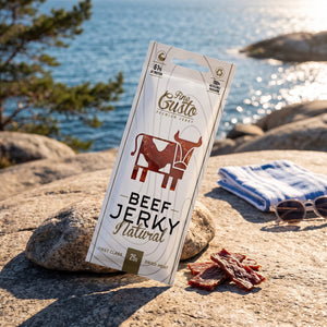 Fine Gusto Beef Jerky Naturell på et svaberg ved sjøen, med håndkle og solbriller, AI-generert bilde