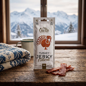 Fine Gusto Kalkun Jerky Naturell på et trebord i en hytte, med ullpledd, emaljekopp og snødekte fjell utenfor vinduet, AI-generert bilde