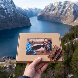 Hånd som holder Indiana Biltong Filet sett med skjærebrett foran et fjordlandskap, AI-generert bilde