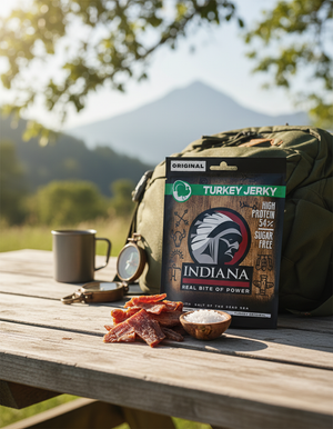 Indiana Kalkun Jerky Original på et bord ute med ryggsekk, kopp og fjell i bakgrunnen, AI-generert bilde