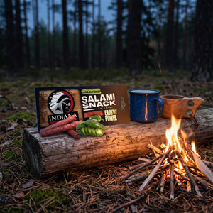 Indiana Salami Jalapeno på en stokk ved bålet i furuskogen i skumringen, med emaljekopp og kuksa, AI-generert bilde