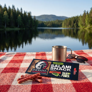 Indiana Salami Jalapeno på et rutete piknikteppe ved et skogsvann en sommerdag, med stålkopp og solbriller, AI-generert bilde