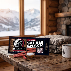 Indiana Salami Original på et trebord i en hytte, med ullpledd, emaljekopp og snødekte fjell utenfor vinduet, AI-generert bilde