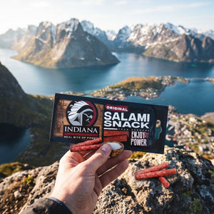 Hånd som holder Indiana Salami Original foran et fjordlandskap, AI-generert bilde