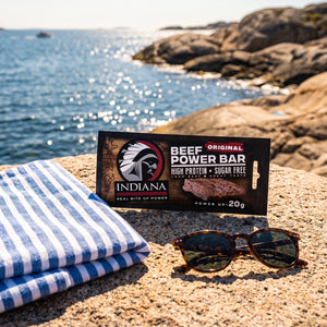 Indiana Beef Bar Original på et svaberg ved sjøen, med håndkle og solbriller, AI-generert bilde