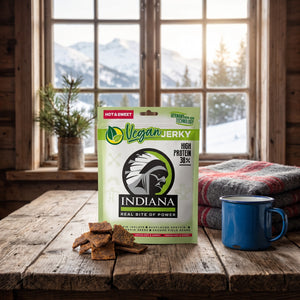 Indiana Vegansk Jerky Hot & Sweet på et trebord i en hytte, med ullpledd, emaljekopp og snødekte fjell utenfor vinduet, AI-generert bilde
