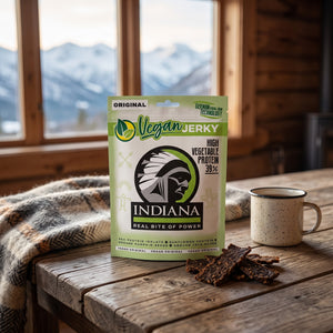 Indiana Vegansk Jerky Original på et trebord i en hytte, med ullpledd, emaljekopp og snødekte fjell utenfor vinduet, AI-generert bilde