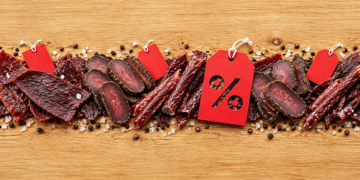 Kjøttsnacks på tilbud: beef jerky, biltong og tørkede pølser på et trebord med røde prislapper