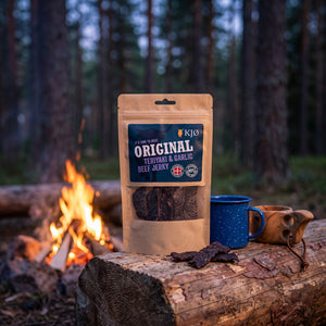 KJØ Teriyaki & Garlic Beef Jerky på en stokk ved bålet i furuskogen i skumringen, med emaljekopp og kuksa, AI-generert bilde