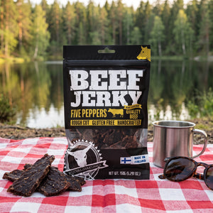 Kuivalihakundi Beef Jerky Fem Pepper på et rutete piknikteppe ved et skogsvann en sommerdag, med stålkopp og solbriller, AI-generert bilde