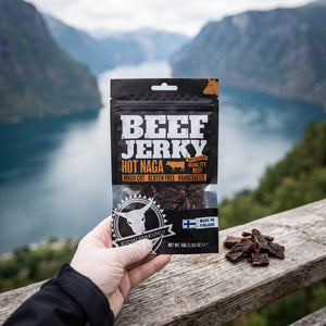 Hånd som holder Kuivalihakundi Beef Jerky Hot Naga foran et fjordlandskap, AI-generert bilde