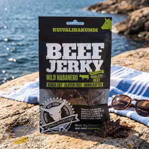 Kuivalihakundi Beef Jerky Mild Habanero på et svaberg ved sjøen, med håndkle og solbriller, AI-generert bilde