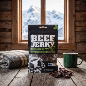 Kuivalihakundi Beef Jerky Mild Habanero på et trebord i en hytte, med ullpledd, emaljekopp og snødekte fjell utenfor vinduet, AI-generert bilde