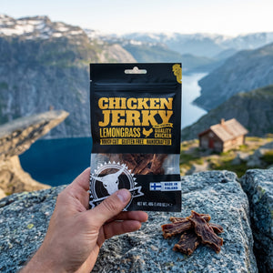 Hånd som holder Kuivalihakundi Kylling Jerky med Sitrongress foran et fjordlandskap, AI-generert bilde