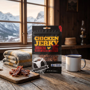 Kuivalihakundi Kylling Jerky Sriracha på et trebord i en hytte, med ullpledd, emaljekopp og snødekte fjell utenfor vinduet, AI-generert bilde