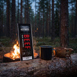 Meat Makers Beef Bar Original på en stokk ved bålet i furuskogen i skumringen, med emaljekopp og kuksa, AI-generert bilde