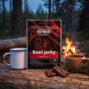Meat Makers Beef Sweet Chilli Gourmet på en stokk ved bålet i furuskogen i skumringen, med emaljekopp og kuksa, AI-generert bilde