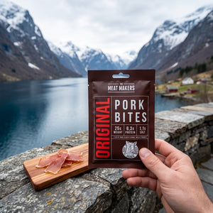 Hånd som holder Meat Makers Lufttørket biter av Svineindrefilet foran et fjordlandskap, AI-generert bilde