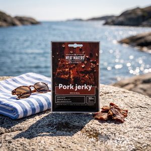 Meat Makers Pork Original Gourmet på et svaberg ved sjøen, med håndkle og solbriller, AI-generert bilde