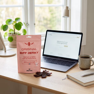 Primal Original Norsk Beef Jerky på skrivebordet ved siden av laptop, kaffekopp og notatbok, AI-generert bilde