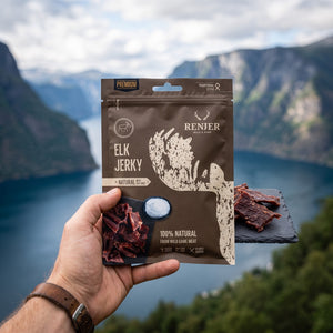 Hånd som holder Renjer Elg Jerky med Sjøsalt foran et fjordlandskap, AI-generert bilde