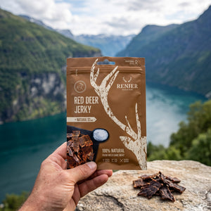 Hånd som holder Renjer Hjort Jerky med Sjøsalt foran et fjordlandskap, AI-generert bilde