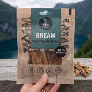 Hånd som holder Sea Snacks tørket brasme foran et fjordlandskap, AI-generert bilde