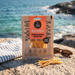 Sea Snacks tørket rødfisk fra Nord-Atlanteren på et svaberg ved sjøen, med håndkle og solbriller, AI-generert bilde