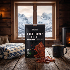 Meat Makers Tørket Kalkun Filet på et trebord i en hytte, med ullpledd, emaljekopp og snødekte fjell utenfor vinduet, AI-generert bilde