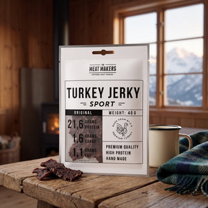 Meat Makers Sports Kalkun Original på et trebord i en hytte, med ullpledd, emaljekopp og snødekte fjell utenfor vinduet, AI-generert bilde